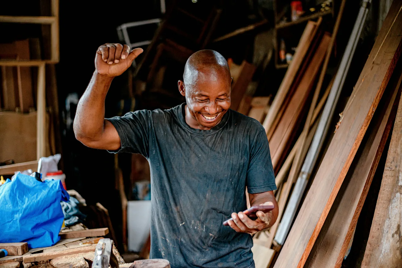 Inhaber eines Handwerksbetriebs steht entspannt in seiner Werkstatt und schaut aufs Smartphone — Feierabend.